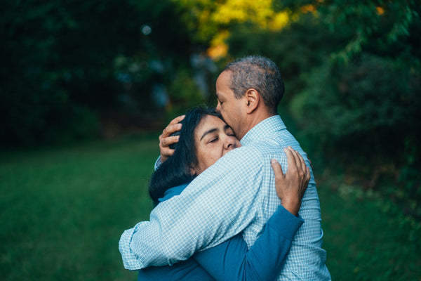 A man kisses his partner on the forehead as they hug under the trees