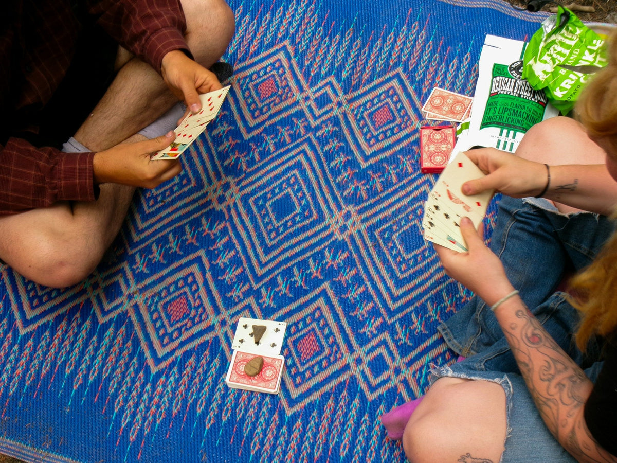 Two people sit on a patterned rug playing a card game, seen from above