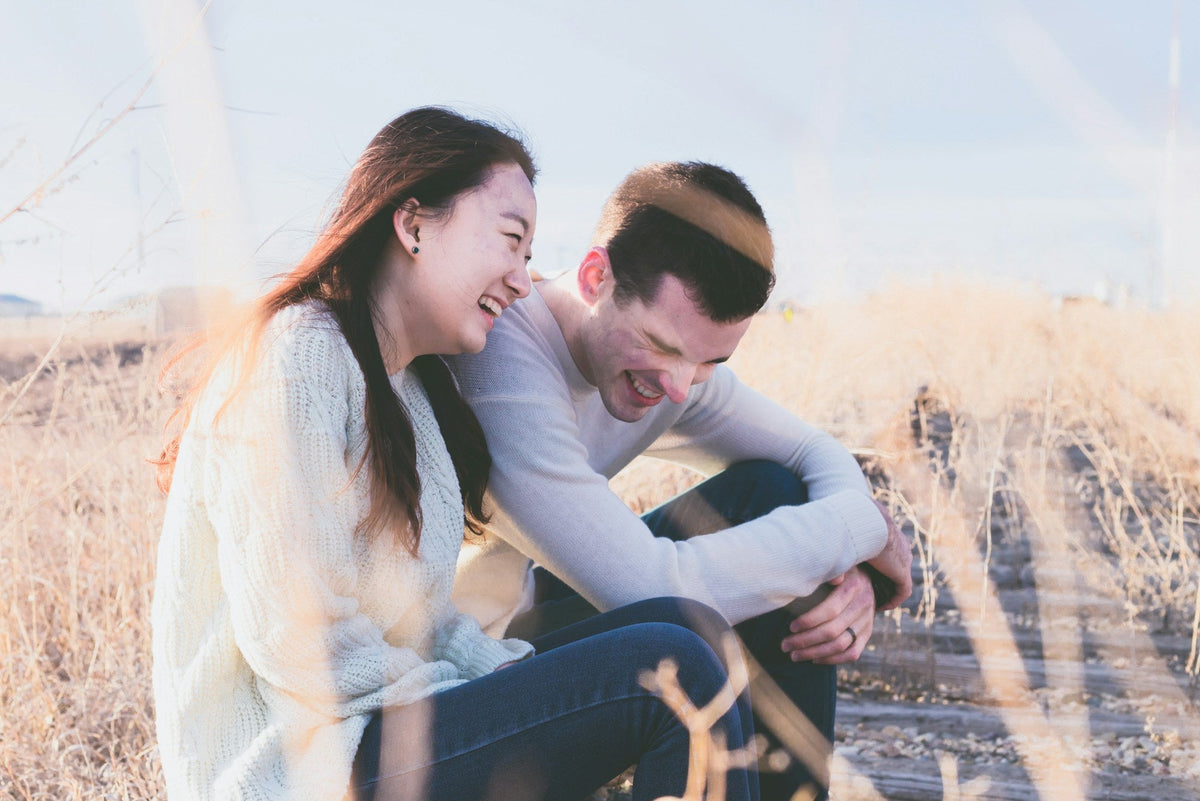 A couple sit close together in long grass, both laughing at something one of them said