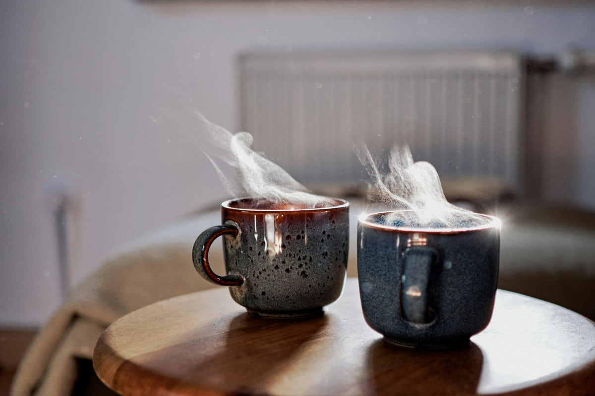 Two steaming speckled mugs side by side on a small wooden table in a quiet, softly lit room